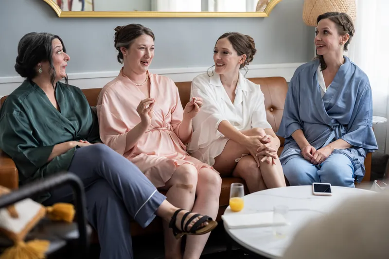 Four women sit together on a couch in the bridal suite, laughing and chatting in casual robes and clothing.