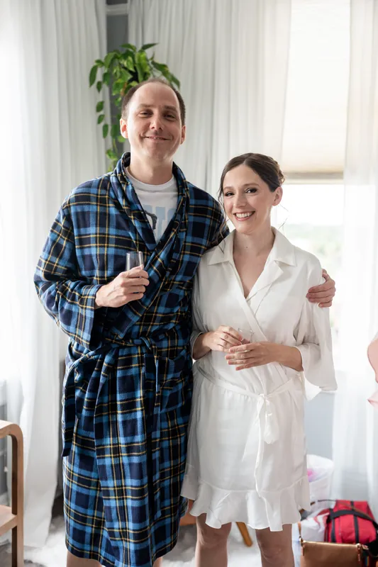 A man in a blue plaid robe stands with his arm around Katherine, who wears a white shirt, both smiling in the getting-ready