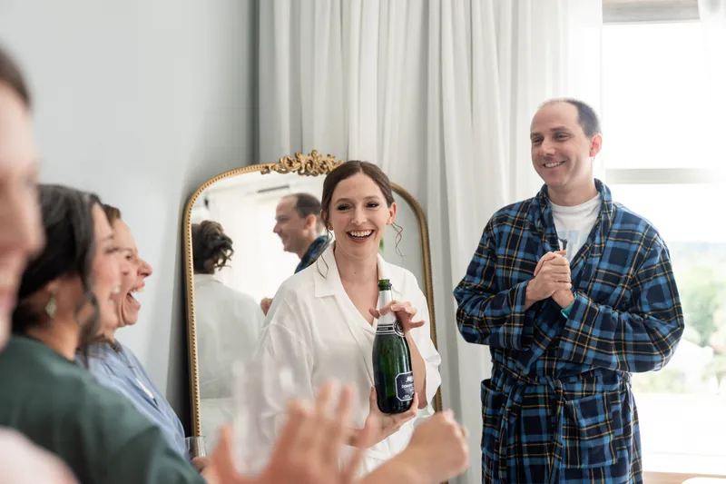 Katherine grins at the camera holding champagne as Nathan raises his glass during a toast with guests around them.