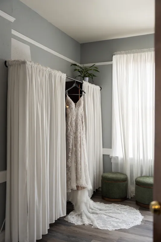A white lace wedding dress hangs on a curtain rod beside sheer white drapes in a gray-walled dressing room.