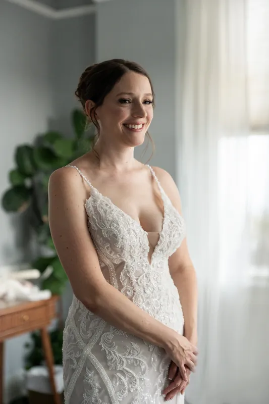 Katherine stands by a window in her beaded lace gown, smiling softly at The Treasury on the Plaza.