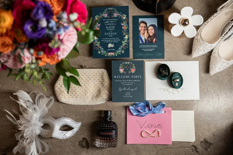 Wedding stationery, ivory heels, colorful flowers, and dark green ring boxes arranged on a neutral linen surface.