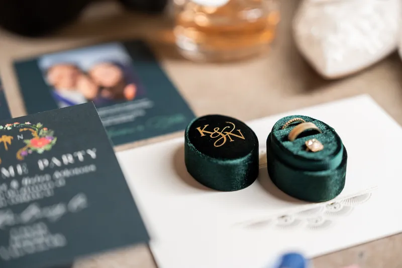 Two dark green velvet ring boxes with gold initials sit on white silk beside navy wedding programs.