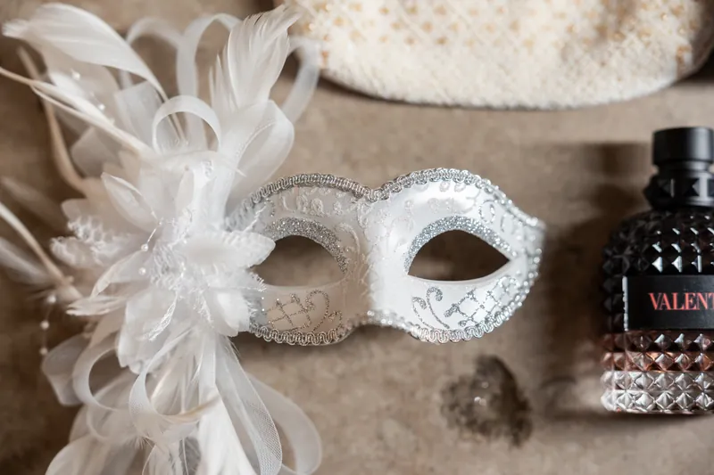 A white sequined masquerade mask with feathered details rests on neutral fabric next to a dark glass bottle.