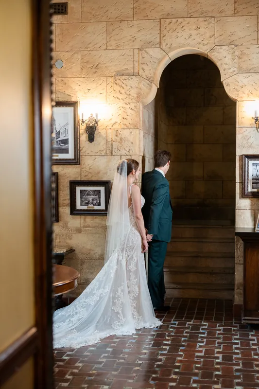 Katherine and Nathan stand facing an arched stone doorway in period costume, her lace train trailing behind them.