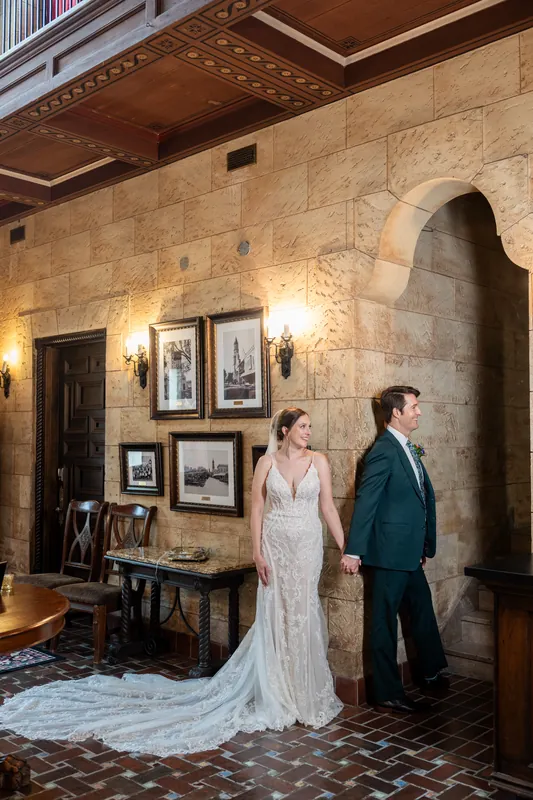 Katherine in a beaded wedding gown and Nathan in a teal suit stand together in the stone-walled venue, her train pooling