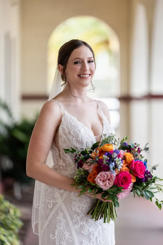 Katherine holds a vibrant bouquet of pink, red, orange, and purple flowers while smiling in the colonnade at The Treasury