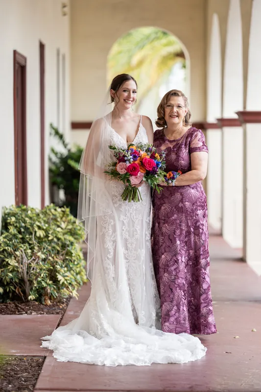 Katherine in an ivory lace gown holds a vibrant pink and red bouquet while posing with a woman in purple at The Treasury