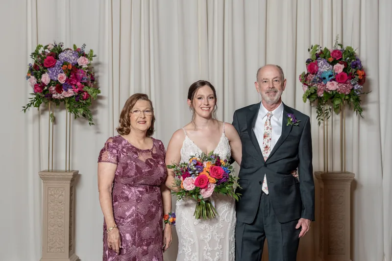 Katherine in white lace stands between two family members in front of tall floral arrangements flanking cream-colored