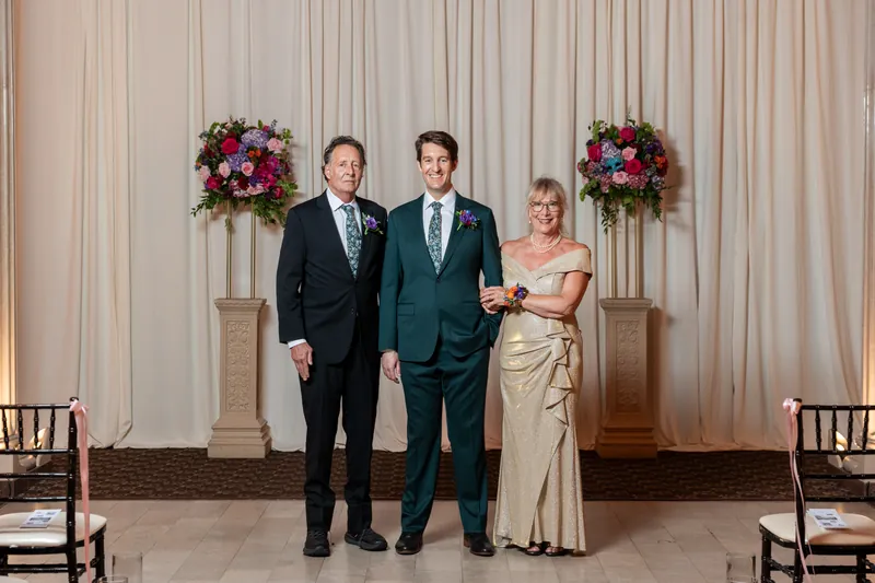 Nathan in a teal suit stands with Katherine and two relatives beneath matching pink and red floral installations