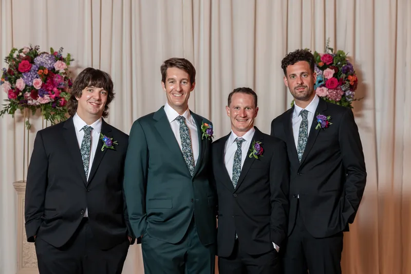 Four groomsmen in dark suits with boutonnières pose together, with one in a standout teal jacket, against draped walls