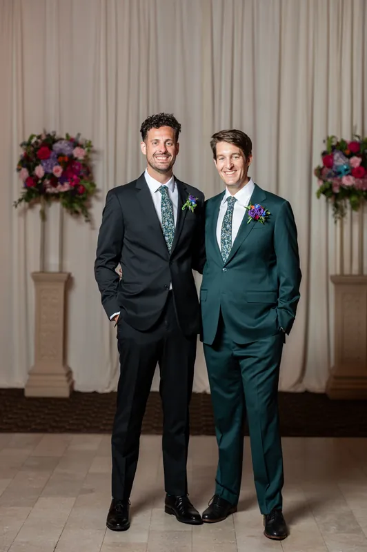 Nathan in a dark suit and his groomsman in teal stand shoulder-to-shoulder, both wearing pink boutonnières, in front