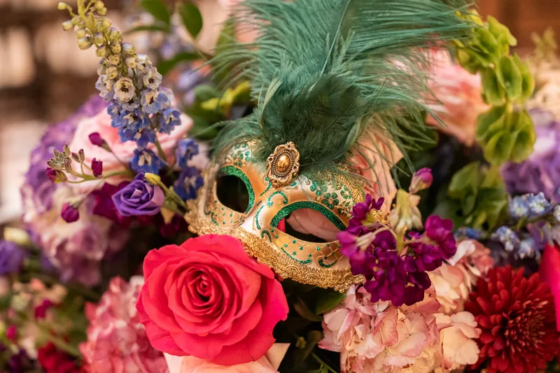 A golden ornate masquerade mask nestled among vibrant roses, peonies, and purple flowers at The Treasury on the Plaza.
