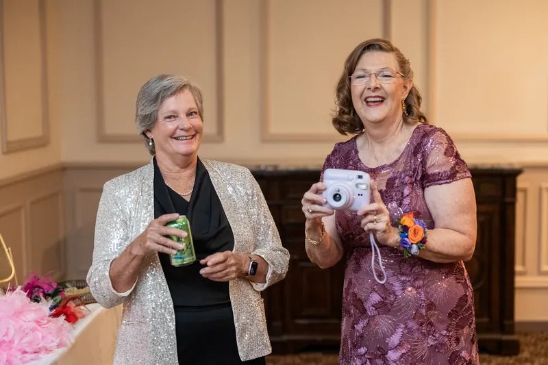 Two women holding cameras smile while standing indoors at The Treasury on the Plaza, with pink floral arrangements visible