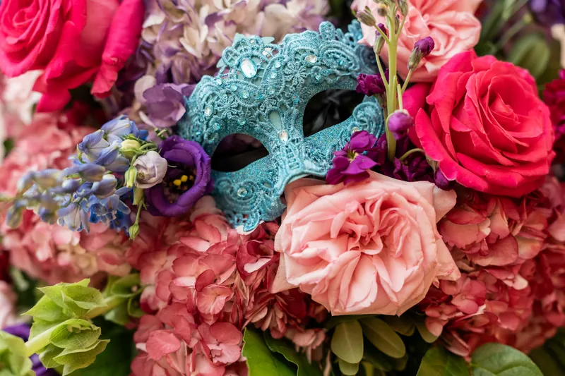 A turquoise glittered masquerade mask surrounded by hot pink roses and blush peonies in an elaborate floral arrangement.
