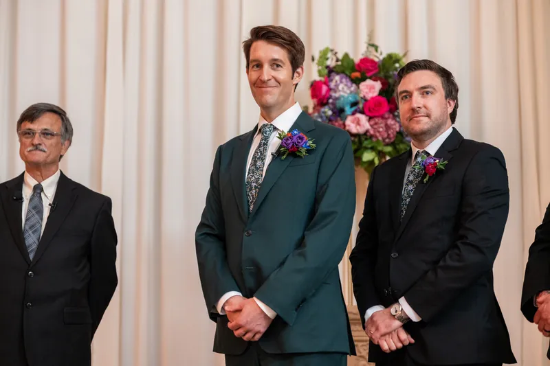 Nathan stands between two groomsmen in dark suits, all wearing boutonnieres, with white curtains and flowers behind them