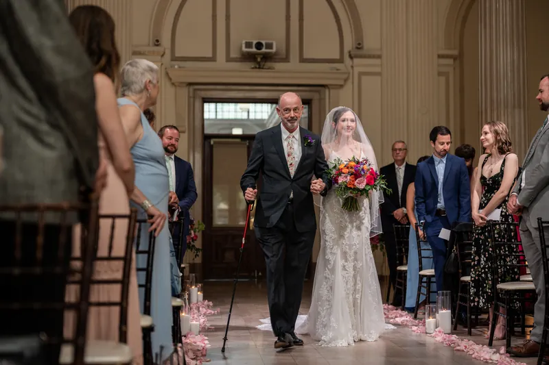 Nathan holds Katherine's hand as they walk back down the aisle after their ceremony, with pink rose petals scattered