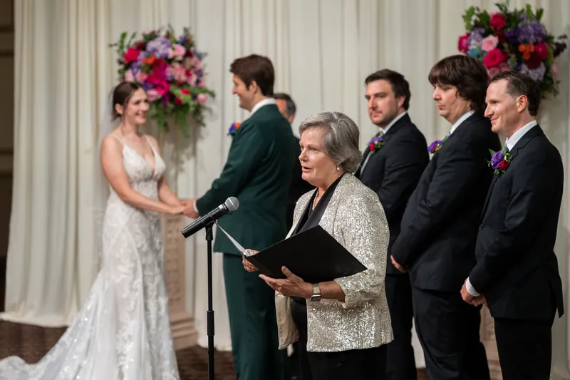 An officiant reads from a book while Katherine and Nathan stand beside each other, flanked by groomsmen and floral