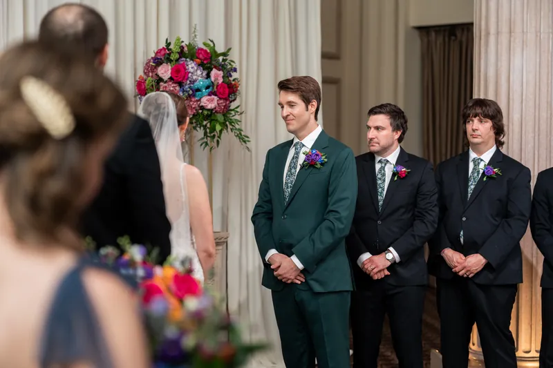 Nathan smiles at Katherine during their ceremony while groomsmen stand nearby and guests look on.
