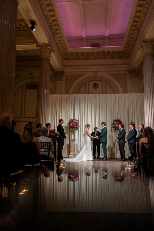 The couple stands beneath soaring columns and pink-lit ceiling as guests look on during their ceremony.