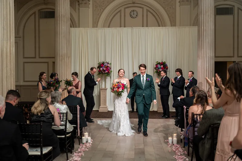 Katherine and Nathan walk back down the aisle hand-in-hand while guests seated on either side celebrate.