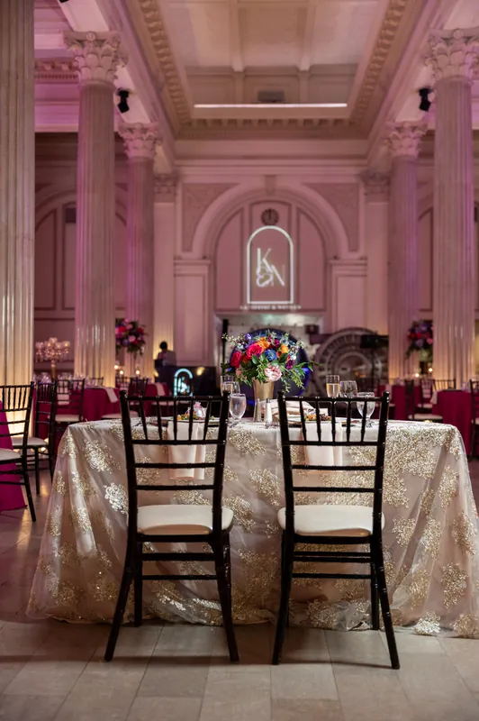 Pale gold sequined linens drape round tables in a pink-lit ballroom with classical columns and a floral arch visible