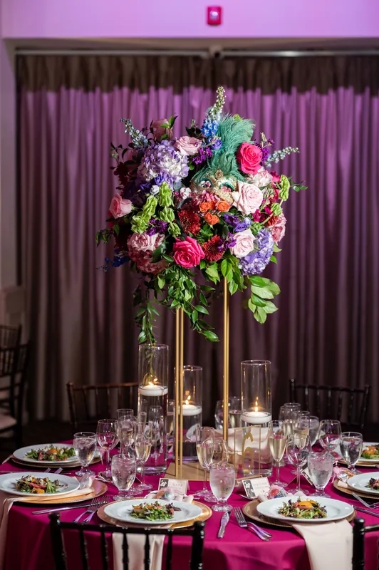 Tall floral arrangement with pink, purple, and white blooms rises from a glass vase above a round banquet table