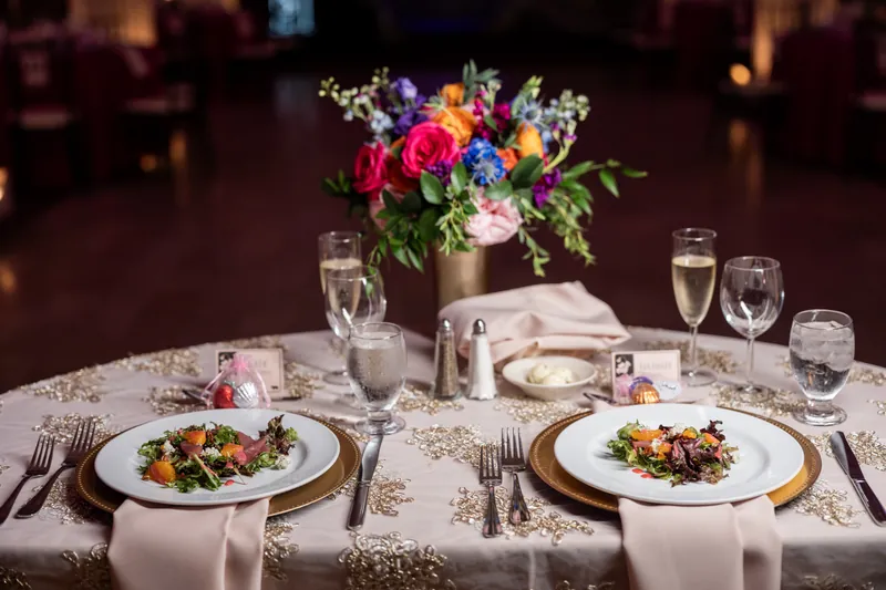 Two place settings with vegetable plates and gold chargers sit across from a low mixed-flower centerpiece on a neutral table