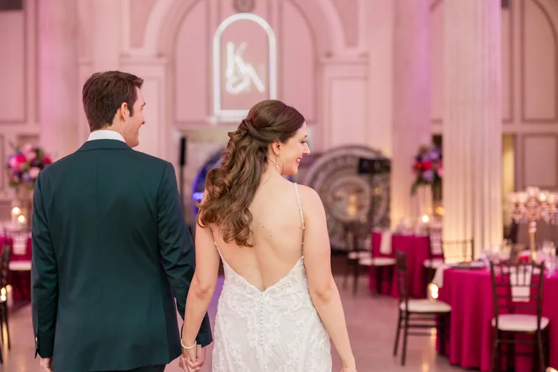 Katherine and Nathan walk hand-in-hand away from the camera through an elegant pink-lit ballroom lined with floral