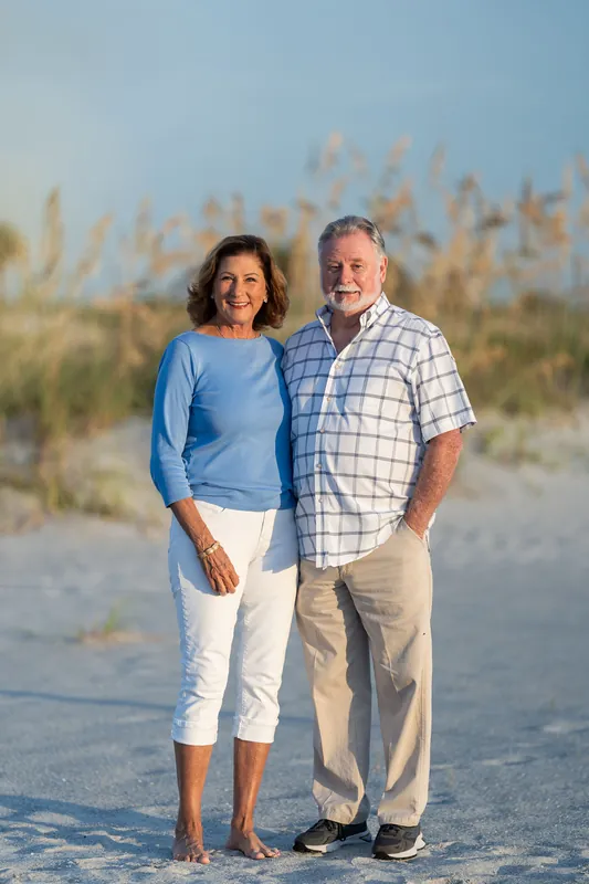 Kelly and a man in casual summer clothing stand together on the sand at Vilano Beach with dune grasses behind them.