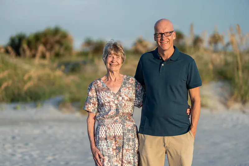 A woman in a patterned dress and a man in a navy polo stand arm-in-arm on the beach at Vilano Beach, dunes visible