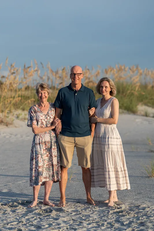 Three people—two women and a man—stand together on the beach at Vilano Beach with dune vegetation behind them.