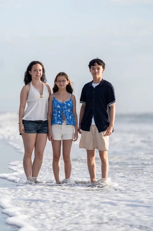 Three young people wade in shallow water at the beach, with the one in the center wearing a blue patterned dress and the two