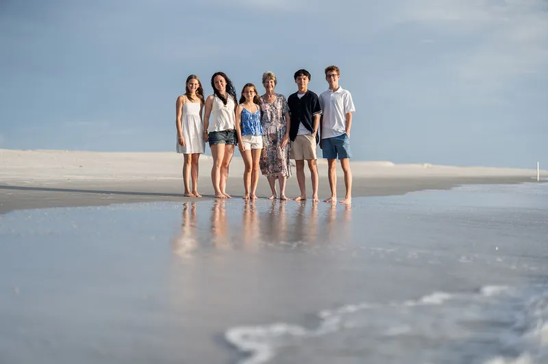 Six family members stand together on the reflective wet sand at the shoreline, with gentle waves visible behind them.