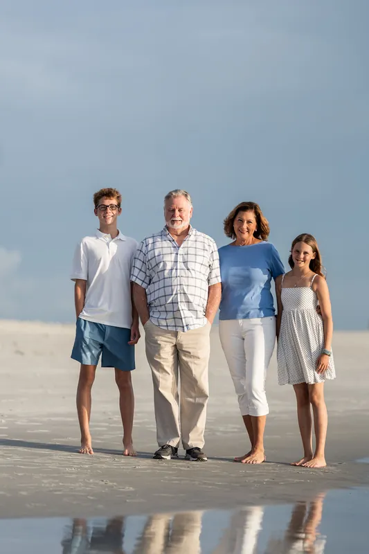 Four people—two teenagers, a middle-aged man, and a young girl—stand barefoot on the beach with their reflections mirrored