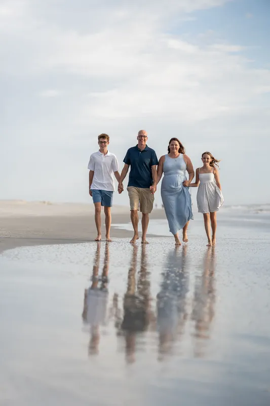 Two adults and two teenagers walk hand-in-hand along the water's edge, their figures reflected in the smooth, wet sand