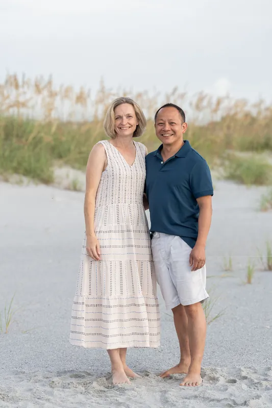 Kelly and a man stand barefoot on the sand at Vilano Beach with dune grasses and shallow water behind them.