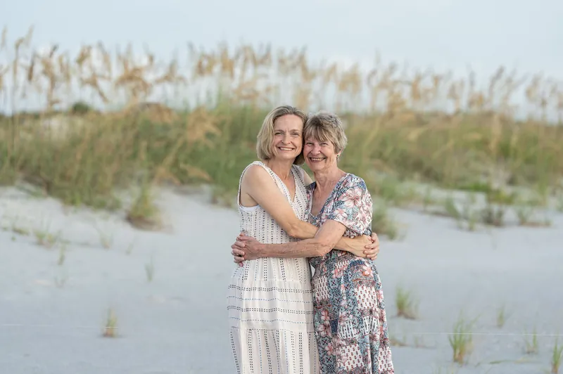 Two women embrace on the beach, both smiling toward the camera with dunes and marsh grass in the background.