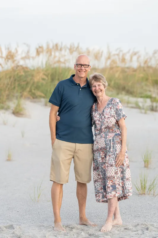 A man in a navy polo and khaki shorts stands beside a woman in a floral dress on the sand at the beach.