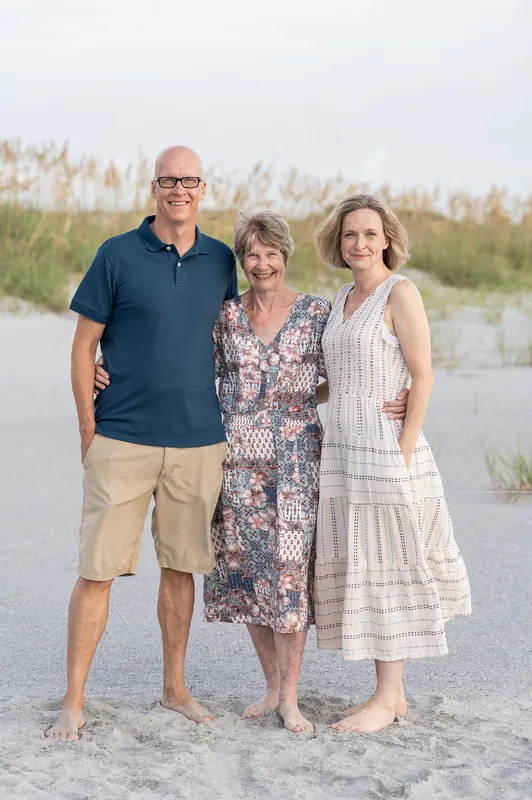 Three people pose together on the beach—a man in navy and khaki, flanked by two women in patterned and striped dresses.