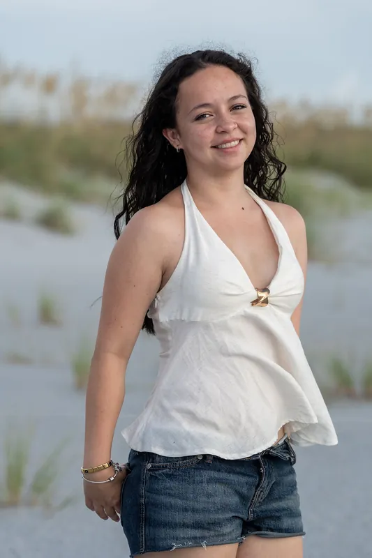 A young woman in a white halter top and denim shorts stands on the beach, smiling with marsh and water visible behind her.