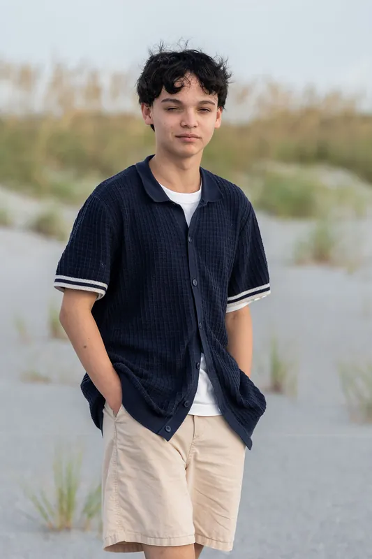 A young man wearing a navy striped shirt and cream shorts stands on the sand with beach grasses and shallow water