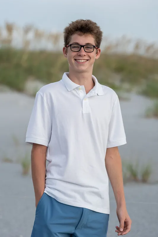 Teen wearing glasses and a white polo shirt stands at Vilano Beach with marsh grasses and water behind him.