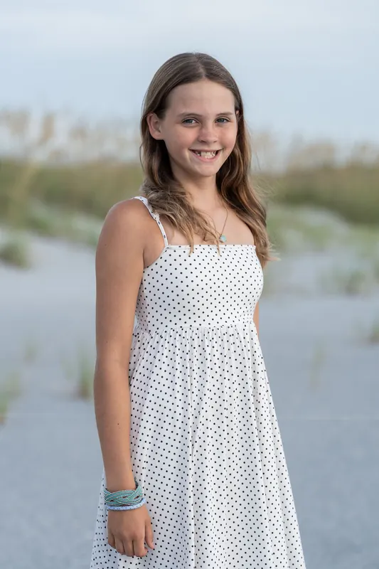 Young woman in a white polka-dot sundress smiles at Vilano Beach with marshland reflected in calm water.