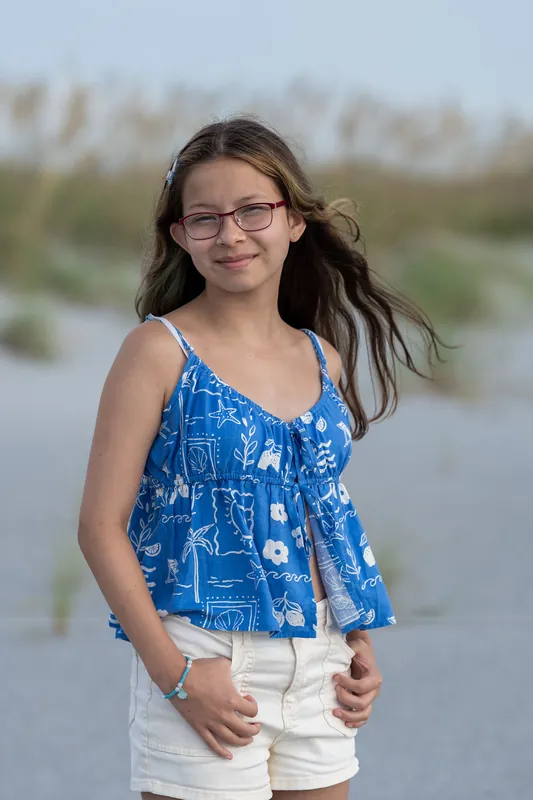 Girl in a blue patterned tank top and white shorts stands at Vilano Beach with soft marsh backdrop.