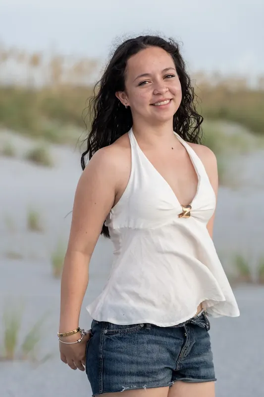A young woman in a white halter top and denim shorts smiles at the camera with the beach behind her.