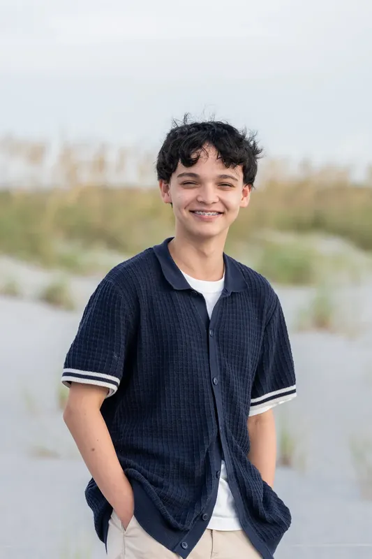 A teenager in a dark striped shirt with white trim stands on the sand, hands in pockets, smiling.