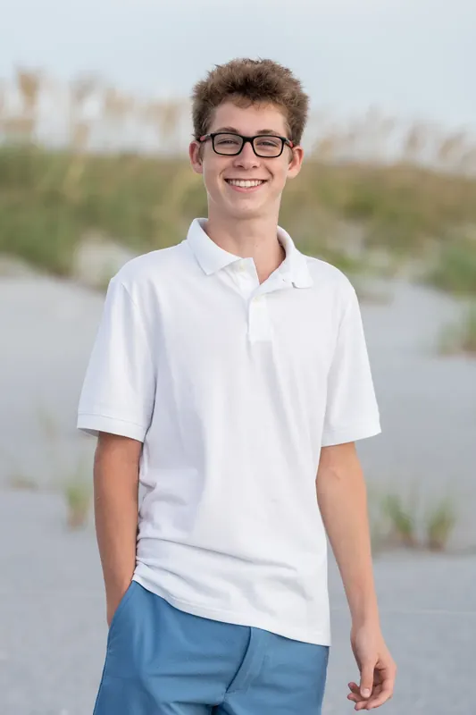 A young man wearing glasses and a white polo shirt poses on the beach with marsh grasses in the background.