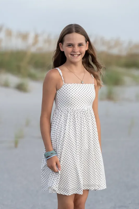 Young person in a white polka-dot dress smiles at Vilano Beach with marsh grasses and water behind them.