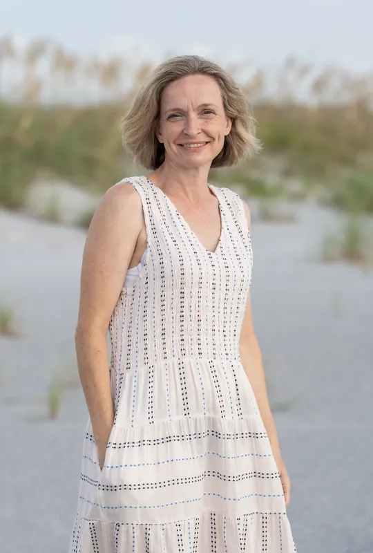 Woman in a white textured sleeveless dress poses with dune vegetation visible along the shoreline.